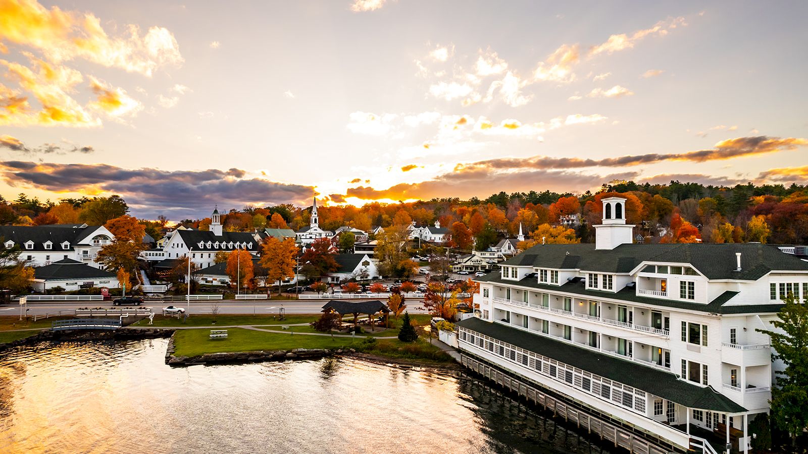 A riverside town at sunset with fall-colored trees and a white multi-story hotel beside the water, glowing in the sky.