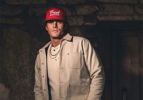 A man wearing a red snapback, beige jacket, and jeans poses with hands near pockets in a dim stone-walled setting.