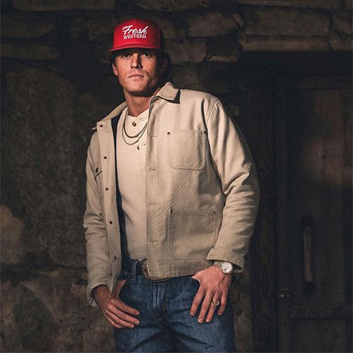A man wearing a red snapback, beige jacket, and jeans poses with hands near pockets in a dim stone-walled setting.