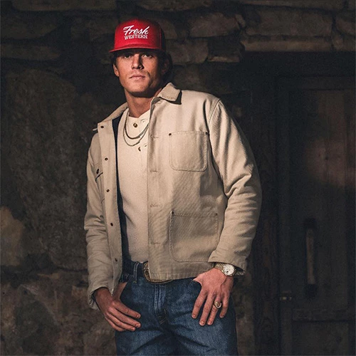A man wearing a red snapback, beige jacket, and jeans poses with hands near pockets in a dim stone-walled setting.