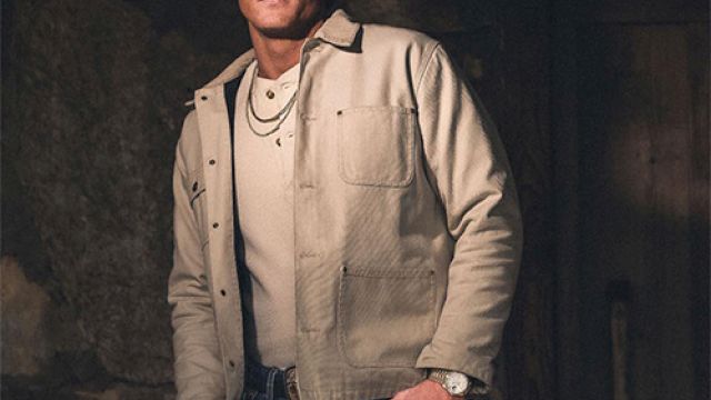 A man wearing a red snapback, beige jacket, and jeans poses with hands near pockets in a dim stone-walled setting.