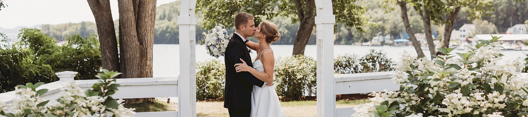 A couple in wedding attire stands kissing under a white arch, surrounded by lush floral bushes in a garden setting by a lake, during daylight.