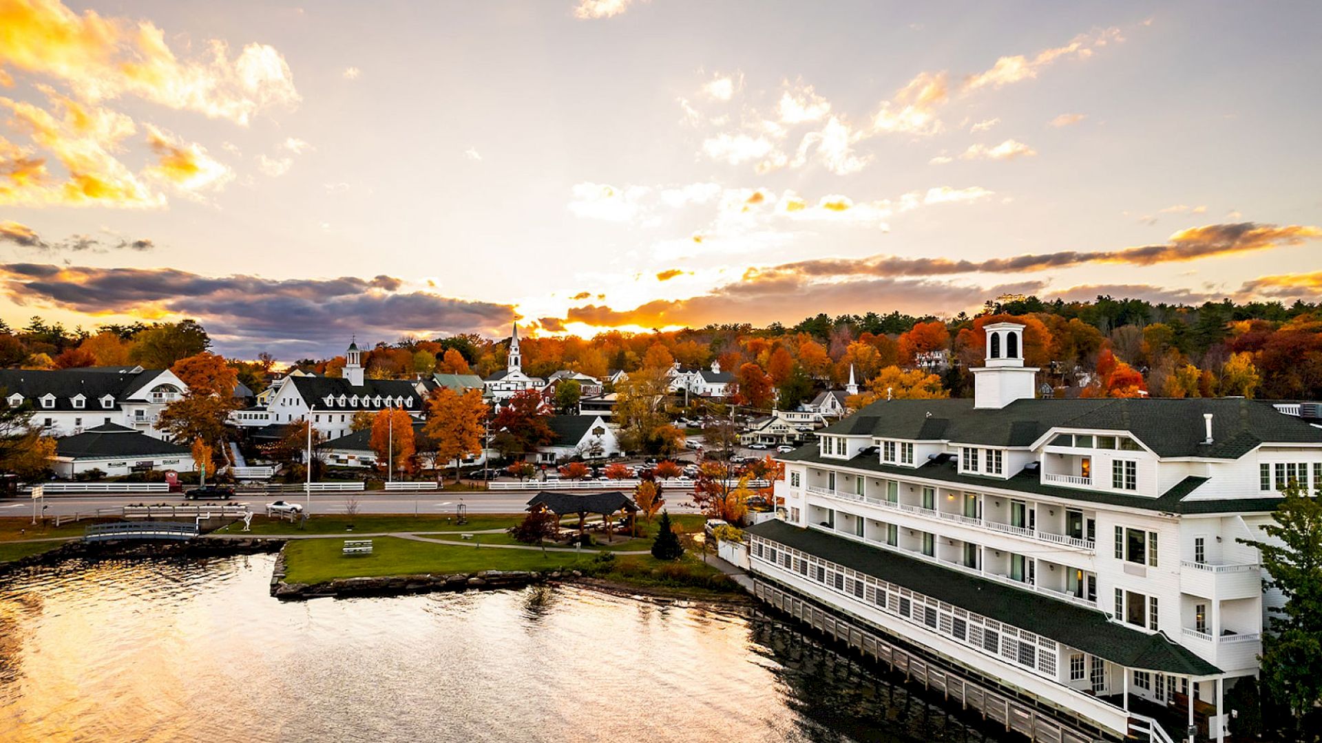 A lakeside town at sunset with white buildings along the shore, colorful autumn trees, and calm water reflecting the sky, ending with a view over the harbor.