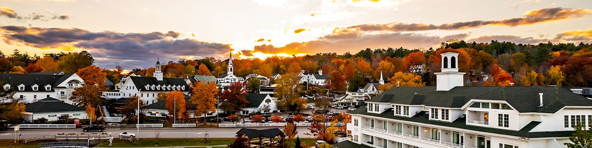 A lakeside town at sunset with white buildings along the shore, colorful autumn trees, and calm water reflecting the sky, ending with a view over the harbor.