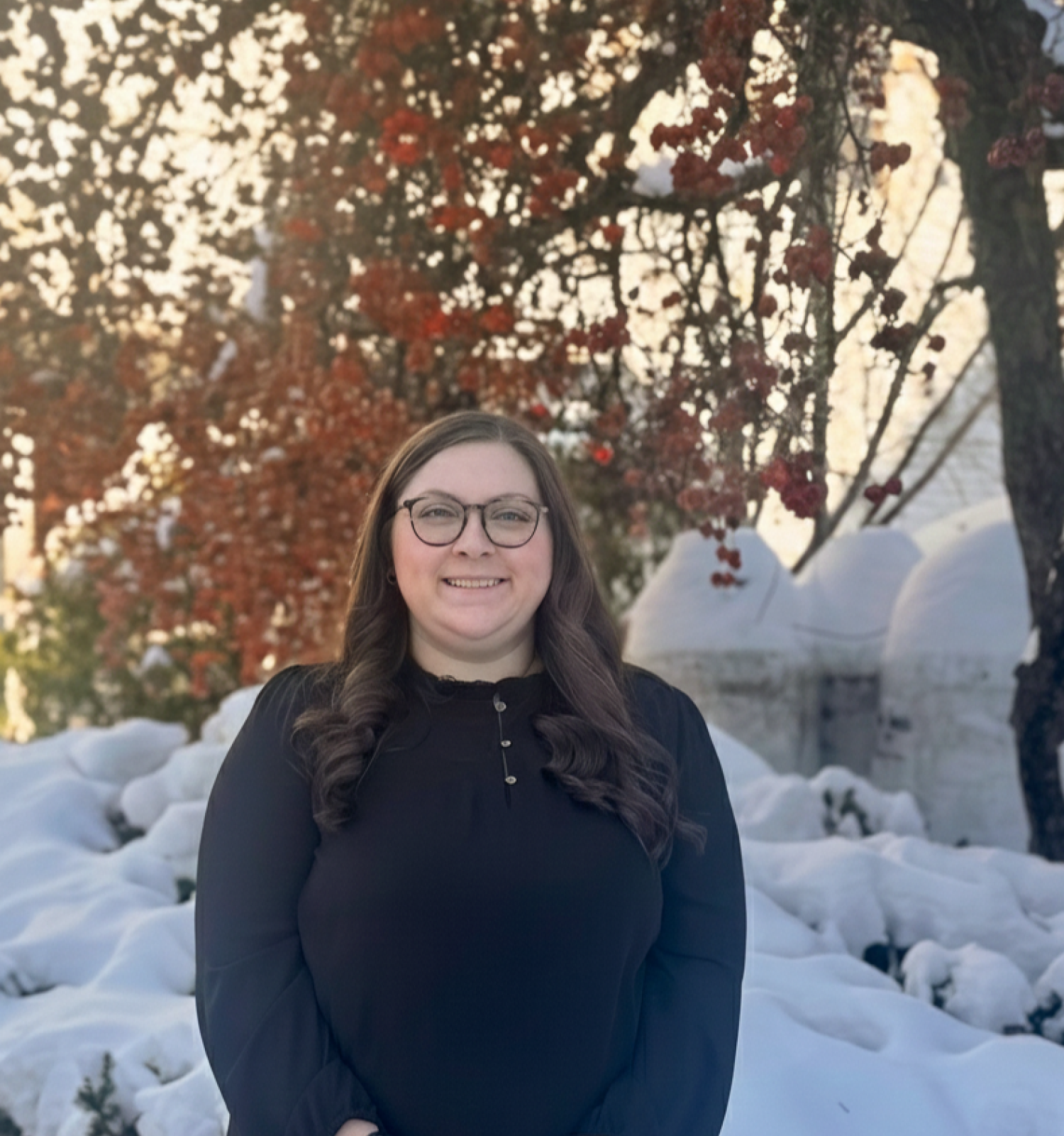 A smiling woman with glasses stands outdoors in a snowy yard, wearing a dark sweater; sunlit trees with red berries glow in the background.