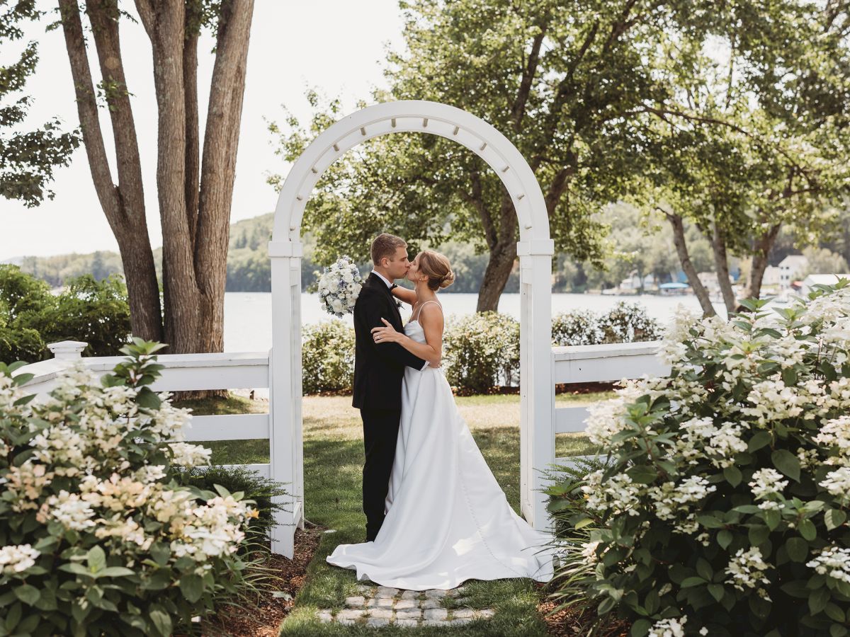 A bride in a white gown and a groom kiss under a white arch, surrounded by lush bushes and trees in a garden setting.