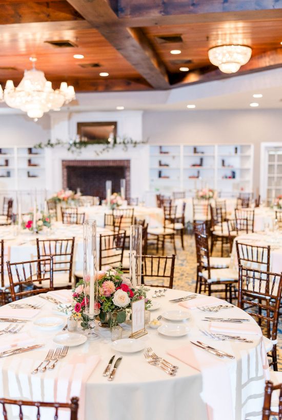 Elegant banquet hall setup with round tables, white linens, floral centerpieces, and wooden chiavari chairs, chandelier lighting, and place settings.