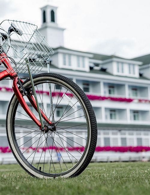 A red bicycle with a front basket stands on a grassy field in front of a white multi-story building with pink accents.