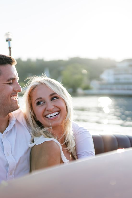 Two smiling people sitting close on a boat by the water, enjoying a sunny day with a distant marina and white buildings in the background.