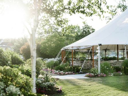 A bright garden scene with a white party tent set up on a lawn, flowering plants, trees, and sunlight streaming through, hinting at a sunny outdoor event.