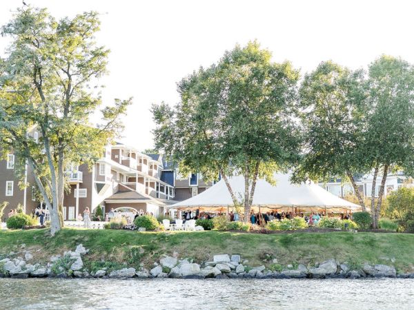 A waterfront scene with trees, a stone-lined bank, and a building with outdoor seating under a white canopy by the water.