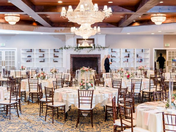 A banquet hall with round tables, ivory tablecloths, chandeliers, and wooden chairs; a fireplace with decor and staff setting up.
