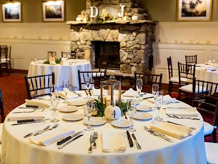 Tables set for a formal dinner in a cozy ballroom with a stone fireplace, candles, and decorative d&eacute;cor. (Ends with a period.)