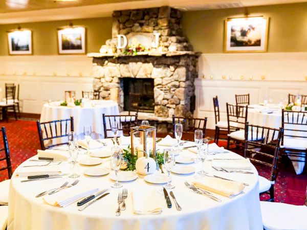 A formal dining room with round tables dressed in white linens, neatly arranged place settings, and a stone fireplace as the centerpiece.
