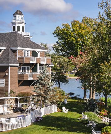 A scenic lakeside view featuring a building with a tower, lush trees, and a grassy area with white chairs and a fence near the water.