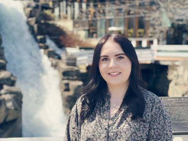 A woman with long dark hair stands in front of a cascading waterfall and a building with large windows, smiling near a deck.