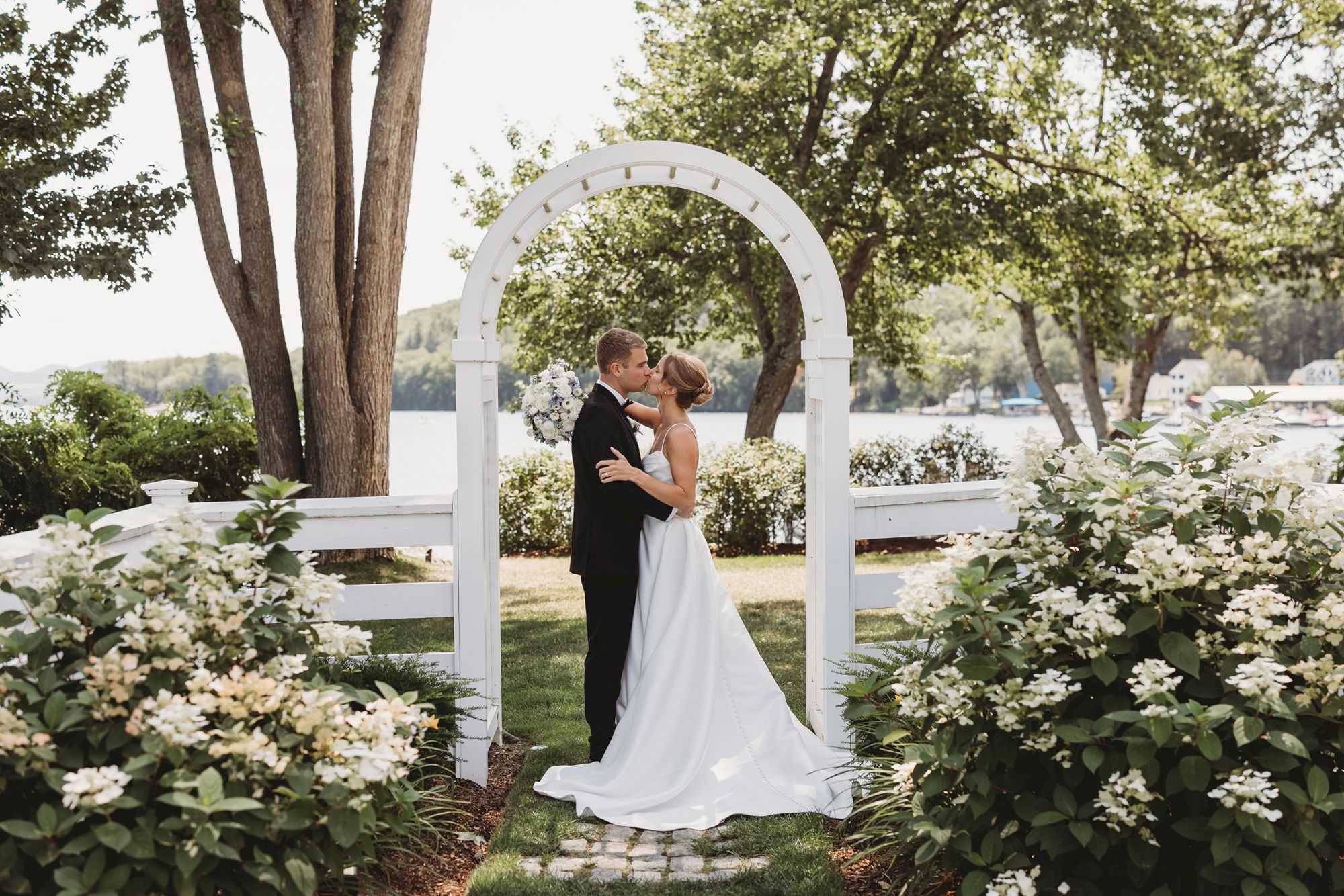 A couple in wedding attire shares a kiss under a white archway, surrounded by blooming bushes in a garden setting.