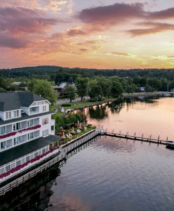 A scenic view of a lakeside building with a dock, water, and a small island at sunset, surrounded by lush greenery and a colorful sky.