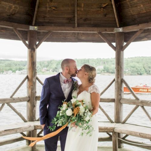 A newlywed couple shares a kiss under a wooden gazebo by the water, with the bride holding a bouquet and the groom in a navy suit.