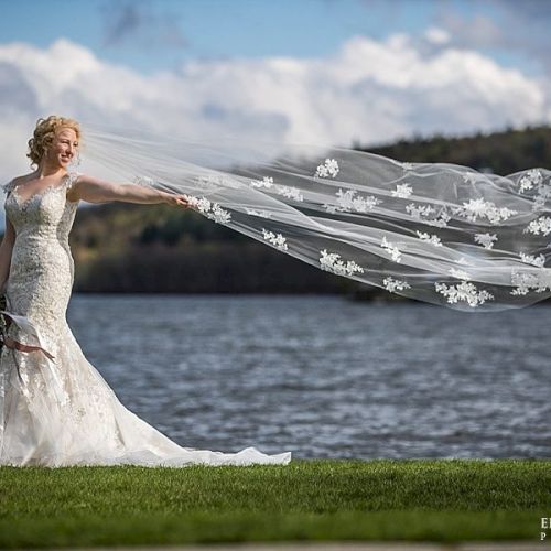 A bride in a white lace gown stands by the water, her long, flowing veil trailing behind her like a delicate ocean breeze.