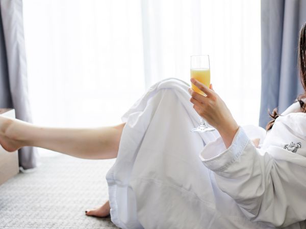 She lounges in a bathrobe on a hotel room floor, holding a drink while one leg stretches out.