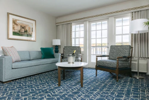 A bright, comfortable living room with a blue patterned rug, light blue sofa, two patterned armchairs, a round coffee table, and large windows letting in natural light. Finishing touches include a floor lamp, framed artwork, and cushions, end result is cozy and airy.