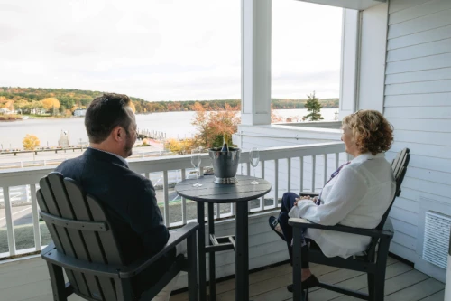 Two people sit on a balcony overlooking a lake in the fall, chatting at a small round table with a bucket of utensils; a peaceful outdoor scene.