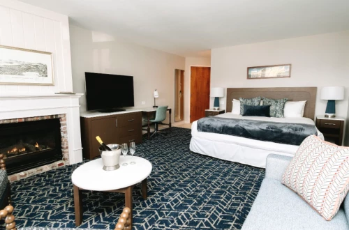 A hotel-style suite with a fireplace, flat-screen TV on a dresser, a king bed with pillows, two nightstands, a sofa, and a patterned carpet.