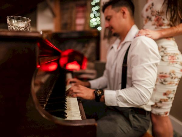 A man playing a piano while a woman stands behind him, as warm lights and a wrapped gift box glow in the background, creating a cozy scene.