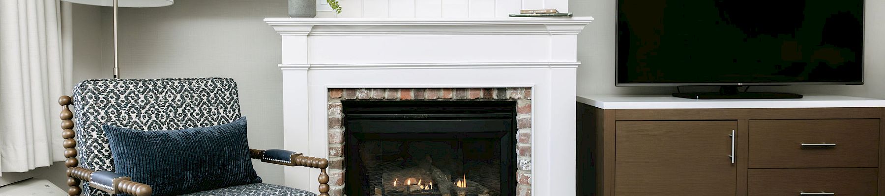 A cozy living room with a fireplace, patterned armchair, blue rug, and a flat-screen TV on a wooden cabinet. The space is bright and neatly styled.