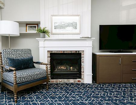 A cozy living room with a fireplace, patterned armchair, blue rug, and a flat-screen TV on a wooden cabinet. The space is bright and neatly styled.