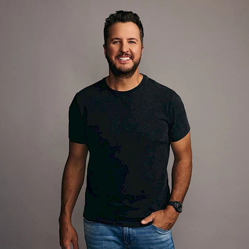 A man with short dark hair and a beard, wearing a black T-shirt and jeans, stands against a gray backdrop with a relaxed smile.