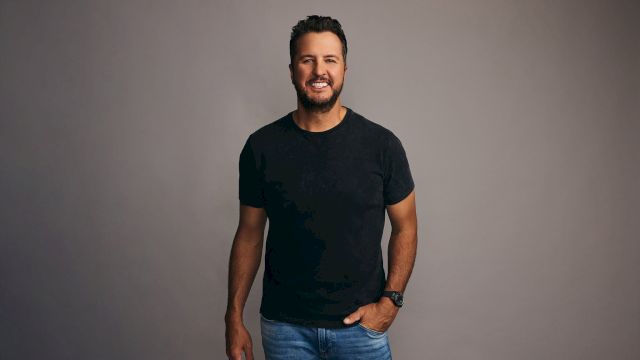 A man with short dark hair and a beard, wearing a black T-shirt and jeans, stands against a gray backdrop with a relaxed smile.