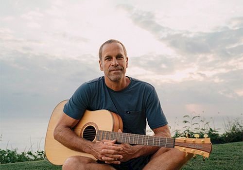 A man sits cross-legged on grass, holding an acoustic guitar, outdoors at sunset, looking calm.