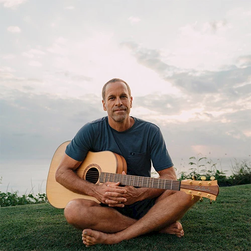 A man sits cross-legged on grass, holding an acoustic guitar, outdoors at sunset, looking calm.