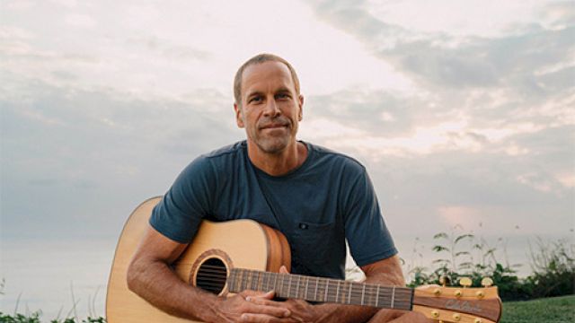 A man sits cross-legged on grass, holding an acoustic guitar, outdoors at sunset, looking calm.