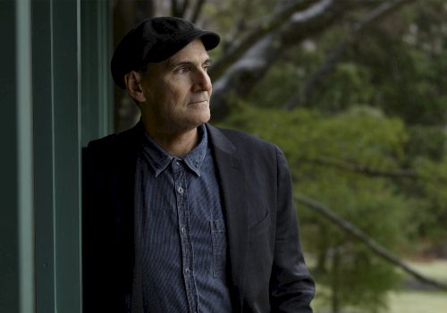 A thoughtful man wearing a dark blazer, denim shirt, and cap stands beside a green wall, gazing into the distance outdoors.