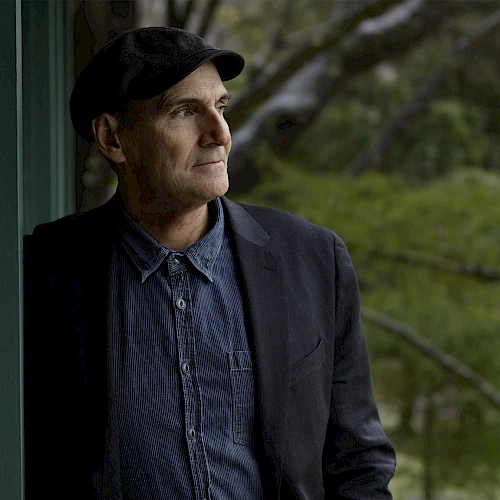 A thoughtful man wearing a dark blazer, denim shirt, and cap stands beside a green wall, gazing into the distance outdoors.
