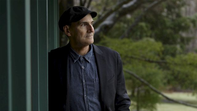 A thoughtful man wearing a dark blazer, denim shirt, and cap stands beside a green wall, gazing into the distance outdoors.