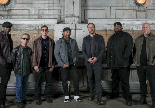 A group of seven men posing for a photo in an industrial setting, wearing casual jackets and dark clothing, standing in a row.