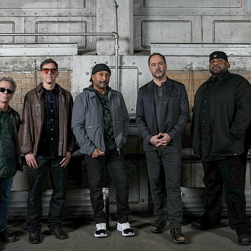 A group of seven men posing for a photo in an industrial setting, wearing casual jackets and dark clothing, standing in a row.