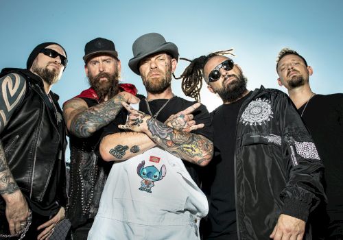 A group of five tattooed men, wearing hats and sunglasses, pose confidently together outdoors, some with arms crossed, under a clear blue sky.