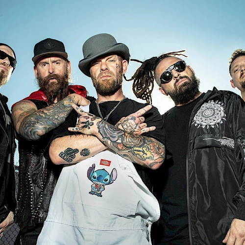 A group of five tattooed men, wearing hats and sunglasses, pose confidently together outdoors, some with arms crossed, under a clear blue sky.