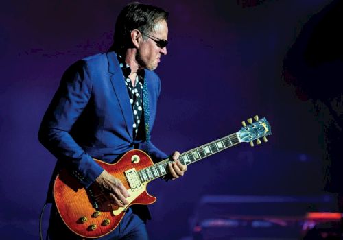 A guitarist in a blue suit and sunglasses plays a sunburst electric guitar on stage.