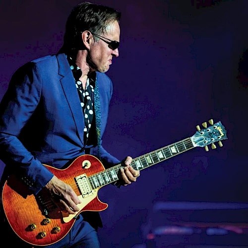 A guitarist in a blue suit and sunglasses plays a sunburst electric guitar on stage.