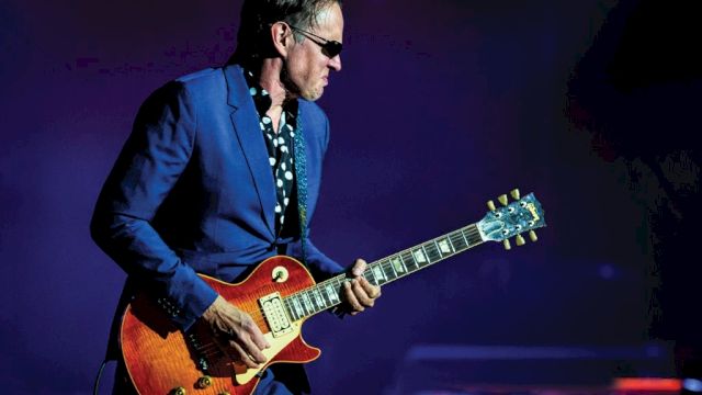 A guitarist in a blue suit and sunglasses plays a sunburst electric guitar on stage.
