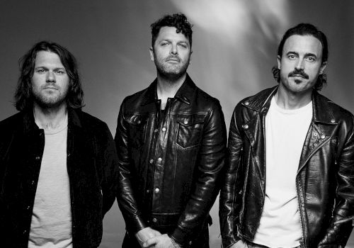 Three men in leather jackets posing for a band photo, black-and-white portrait, serious expressions, casual rock vibe.