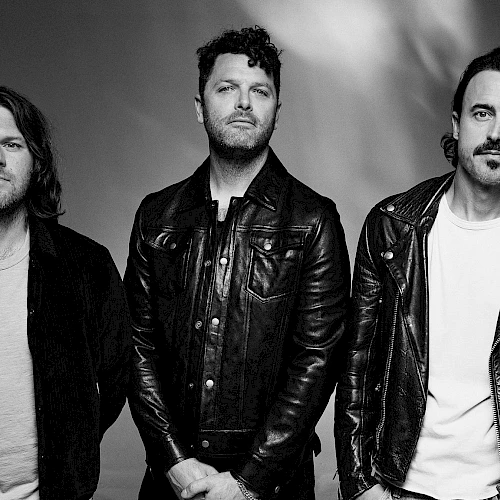 Three men in leather jackets posing for a band photo, black-and-white portrait, serious expressions, casual rock vibe.