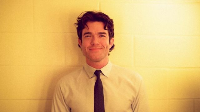 A man with short dark hair in a pale shirt and dark tie stands against a yellow wall, smiling softly at the camera.
