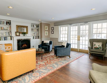 Cozy living room with a fireplace, built-in bookshelves, orange leather sofa, navy chairs, a patterned area rug, and large windows letting in light.